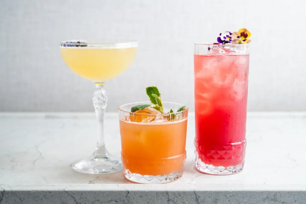 A trio of craft cocktails: a yellow drink in a coupe, an orange drink with mint, and a tall pink drink with a flower garnish