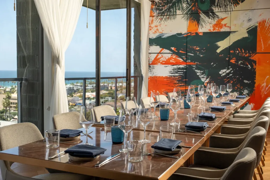 A long, formally set dining table in a private room, featuring a large abstract painting and a floor-to-ceiling window overlooking the coast