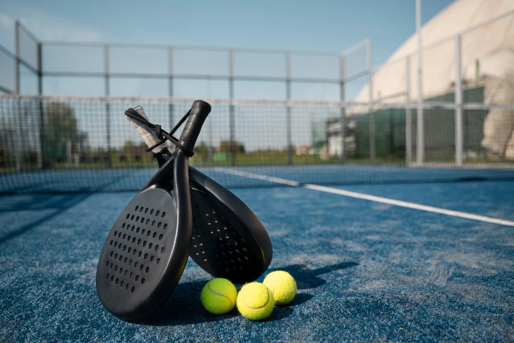 Two black padel rackets and three yellow balls rest on a blue court under a clear sky.