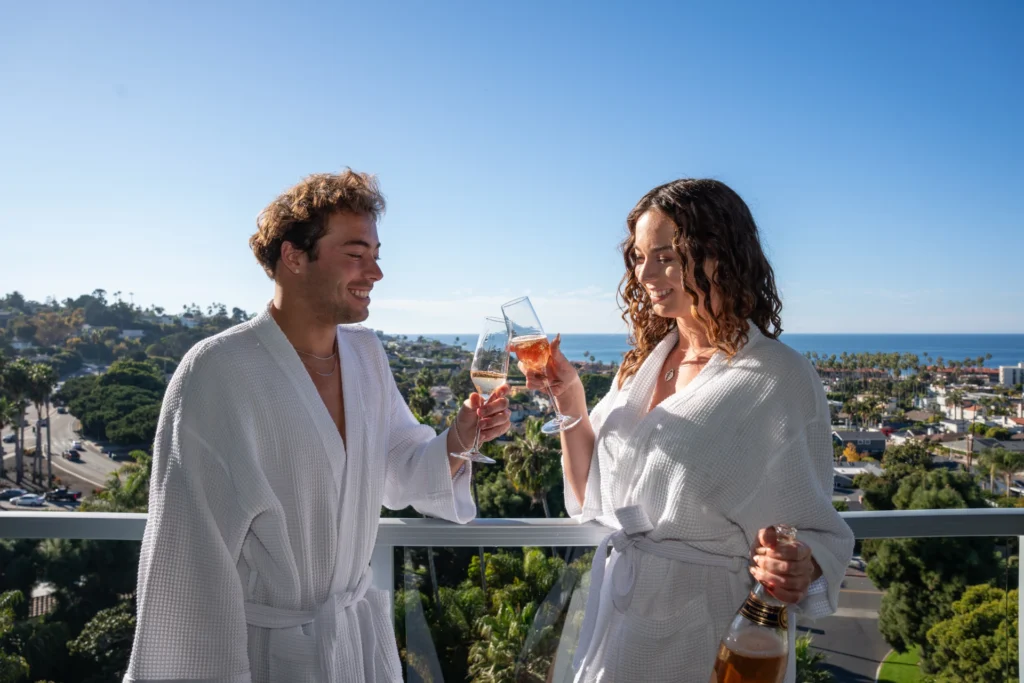 A couple in white robes toasts with champagne on a balcony at luxury hotels in la jolla.