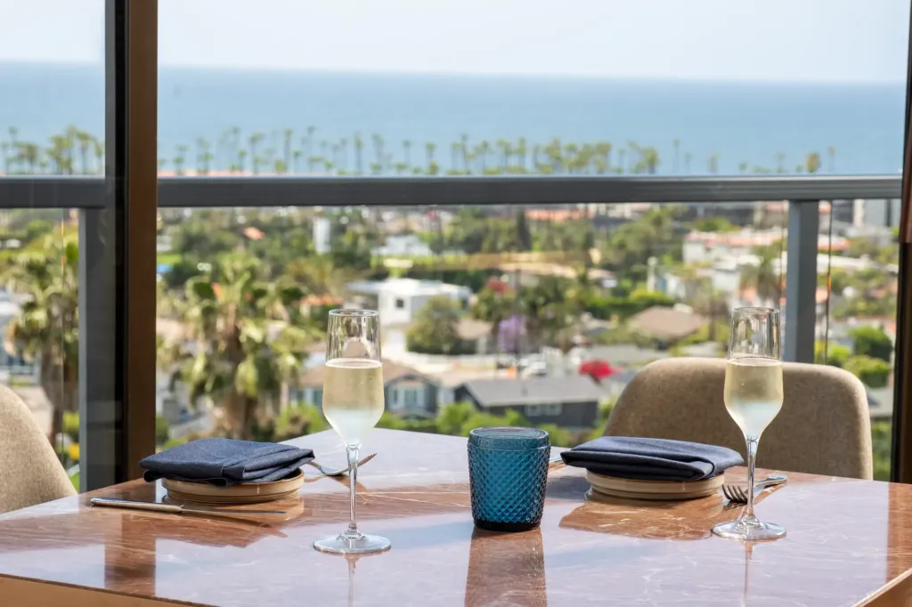 A dining table set for two with flutes of champagne, overlooking a sunny coastal city and the ocean from a large window
