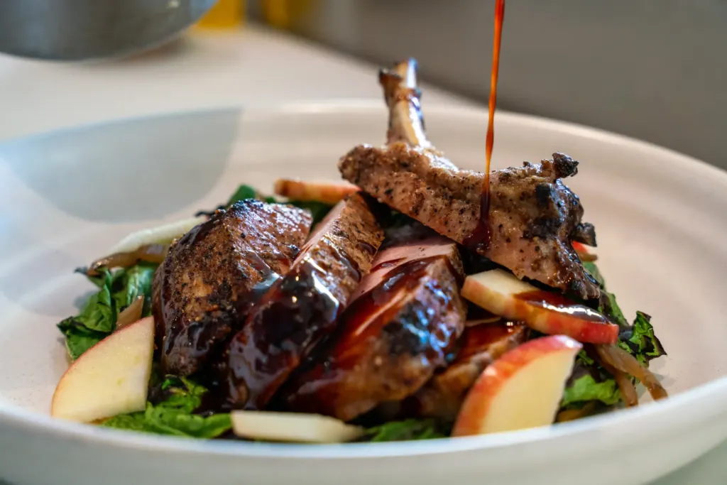 A close-up of a gourmet dish of sliced roasted meat and apples, with a rich, dark sauce being drizzled over the top